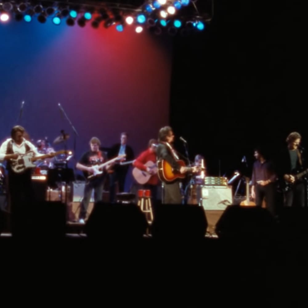 A musical performance at an indoor concert venue with colorful stage lights illuminating the performers on a dark stage setup.
