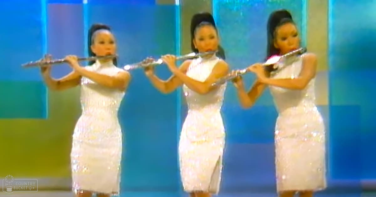 Three young adult female flutists perform indoors against a blue abstract backdrop with bright, even lighting.