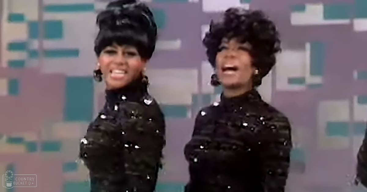 Two adult female performers wearing black sequined tops with long sleeves and high necklines, set against a patterned wall backdrop suggesting a studio or stage environment.