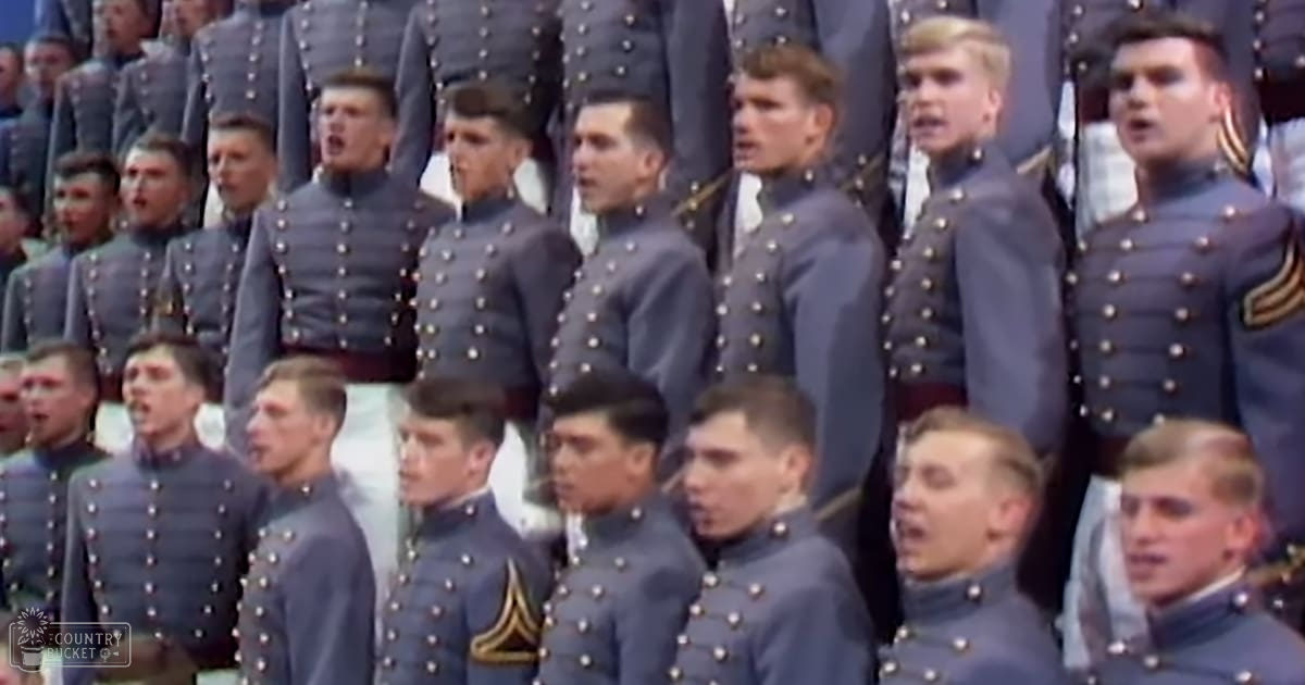 Young adult males in dark blue military-style uniforms with gold trim, outdoors with a crowd behind them.