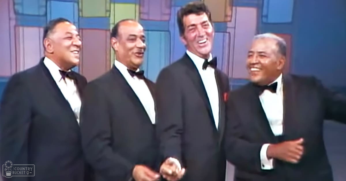 A group performance scene with middle-aged and older adult male performers in formal black tuxedos against a stylized backdrop resembling buildings and windows, set in an indoor stage environment with even artificial lighting.