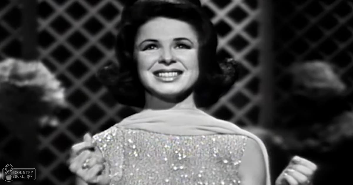 A woman in an adult age band wearing a sleeveless sequined or beaded dress with a fitted cut and flared bottom, likely from the 1950s. The setting includes a lattice structure suggesting a garden or patio area.