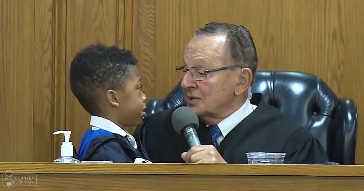 A young boy in a dark-colored robe interacting with an older adult wearing a black judicial robe and glasses within a courtroom setting.