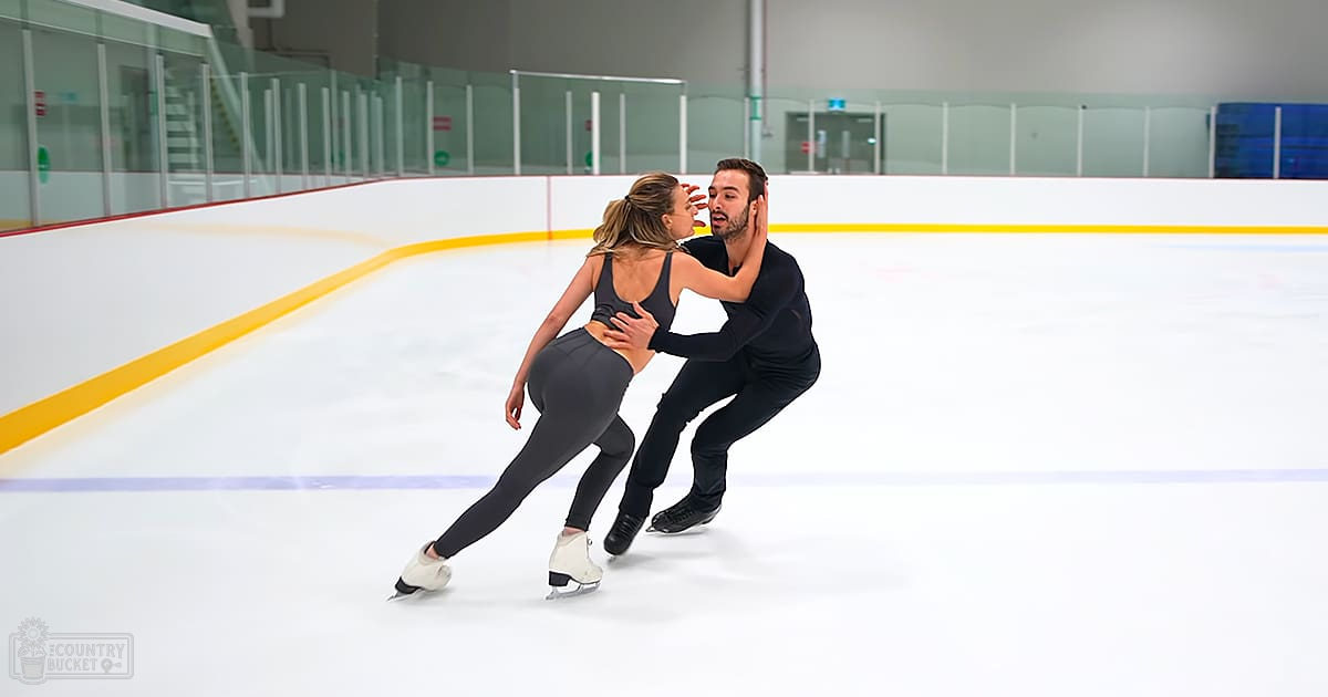 A young adult female figure skater in a black tank top with a scoop neckline and gray leggings, alongside an adult male figure skater wearing a black long-sleeve shirt and black pants, skating on an indoor ice rink with clear glass walls and yellow boundary lines.
