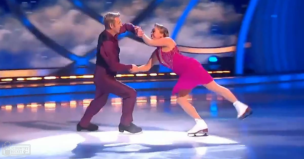A male figure skater in a middle-aged to older adult appearance, dressed in a formal dark maroon outfit and black ice skates, performs alongside a young adult female figure skater wearing a bright pink fitted dress with white ice skates. The performance takes place indoors in an arena lit by blue stage lights against a backdrop resembling clouds or abstract shapes.