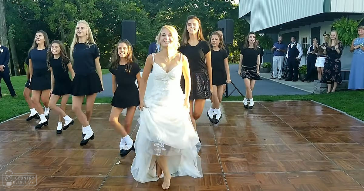 A young adult female bride wearing a white satin wedding gown at an outdoor wedding reception with teenagers and adults in formal attire, surrounded by a wooden dance floor and speakers.