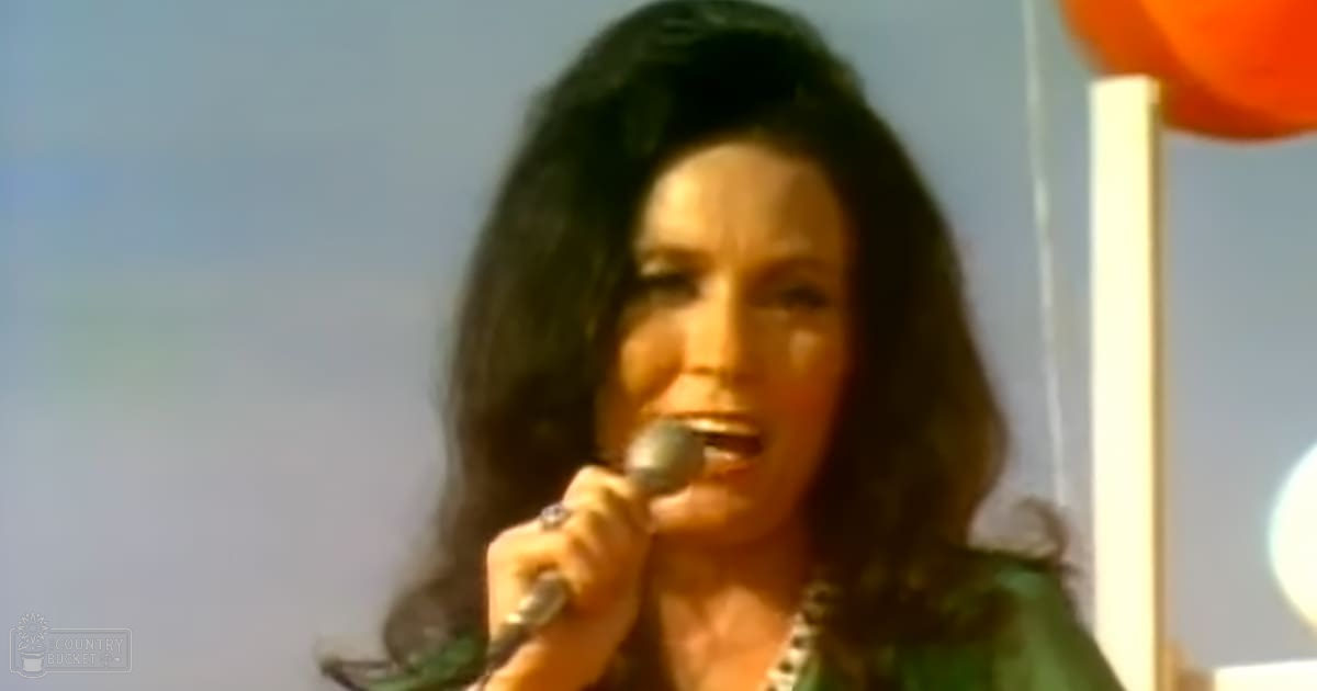 A middle-aged female singer in a casual yet stylish 1970s or early 1980s outfit, performing into a microphone at an outdoor spring or summer event.