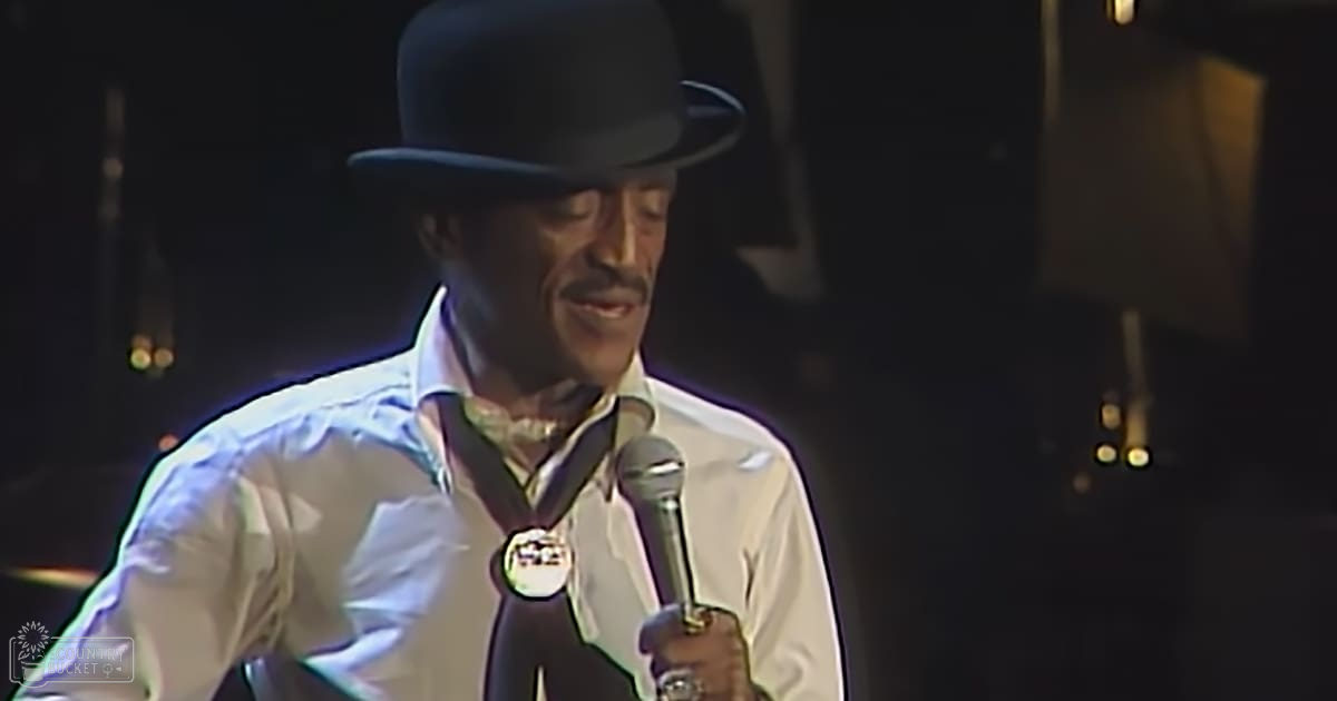 A middle-aged male performer in a white dress shirt and black bow tie stands on stage with a microphone and medal around his neck, suggesting a formal performance setting.