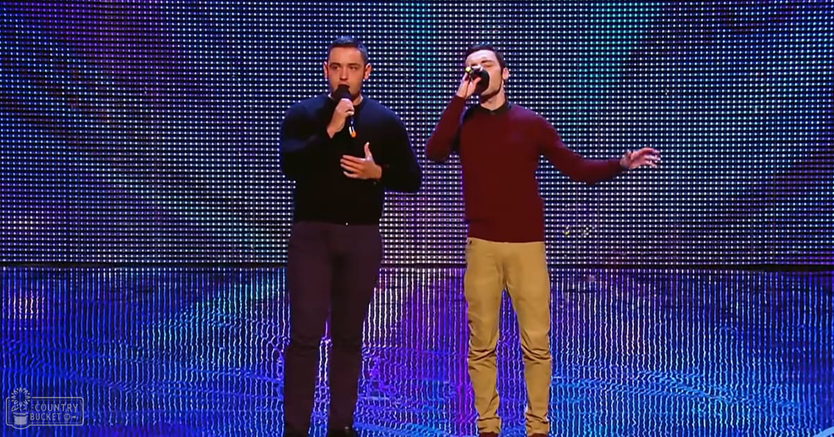 Two adult male performers on stage during a concert or performance in an indoor setting with LED screens displaying blue and white patterns.