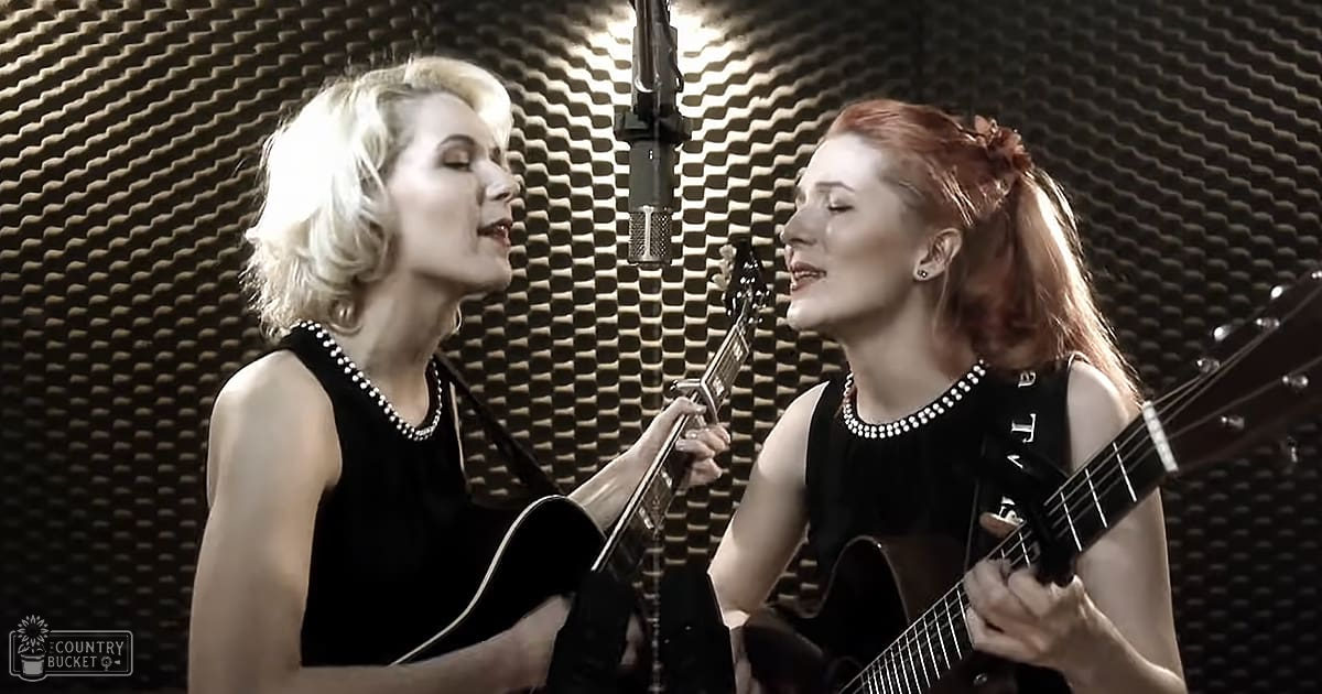 Two adult female singers wearing sleeveless black dresses and pearl necklaces, performing with acoustic guitars in a recording studio setting.