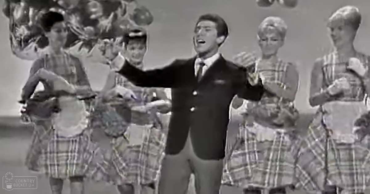 An adult male singer performing on stage with young dancers in a plaid skirt theme.