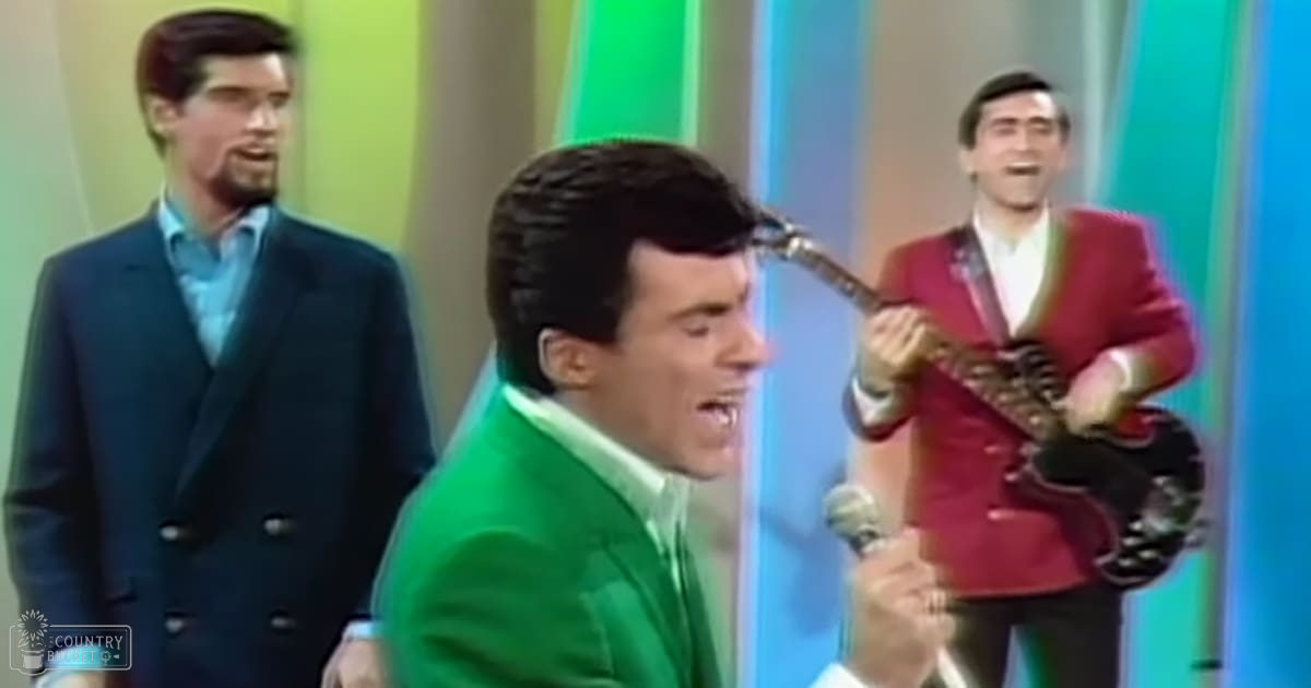 A male adult singer in a bright green suit jacket performs into a microphone with a colorful striped backdrop, accompanied by two other men playing instruments.