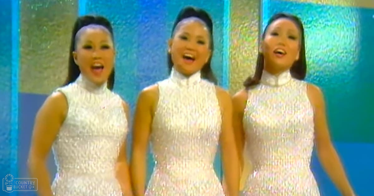 Young adult female performers in a retro-style studio setting, wearing sleeveless high-necked white dresses with sequin-like texture against a blue textured wall.