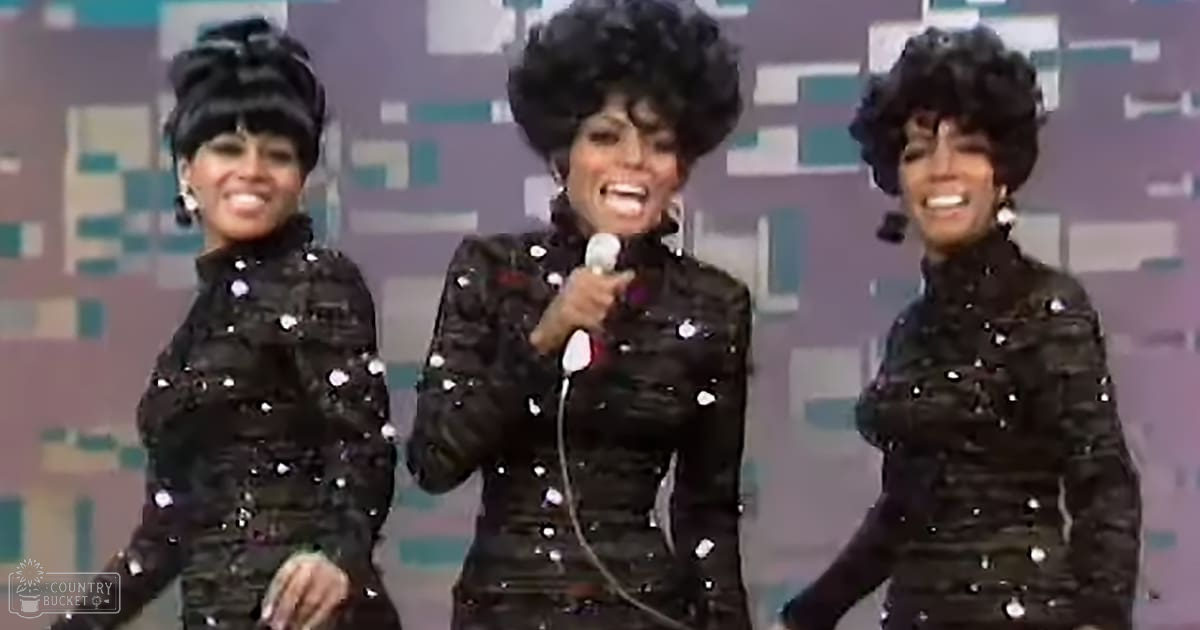 A performance group from the 1960s or 1970s on stage with adult females wearing black sequined outfits and large hoop earrings, holding a microphone against a backdrop of muted geometric patterns.