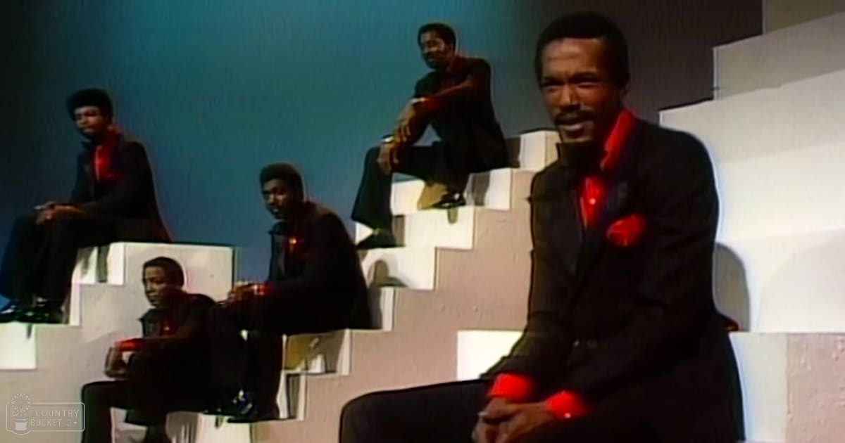 A group of men in dark suits with red accents, seated indoors against a light blue backdrop under artificial lighting.