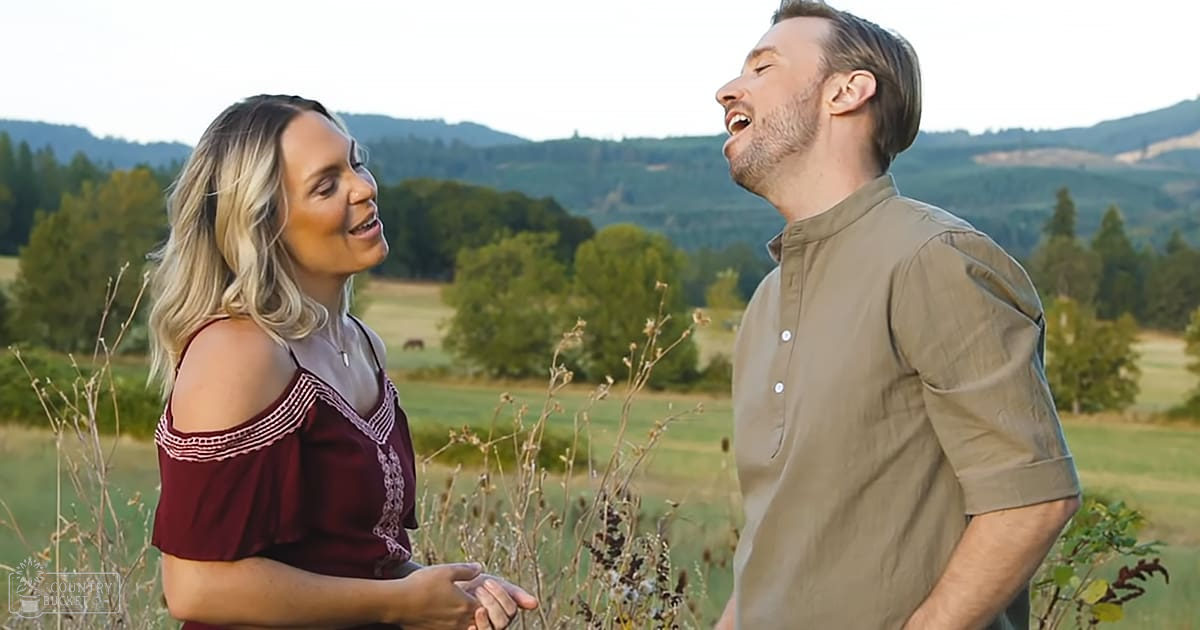 A couple in an outdoor rural setting, likely late summer or early fall with soft golden lighting and greenery around them.