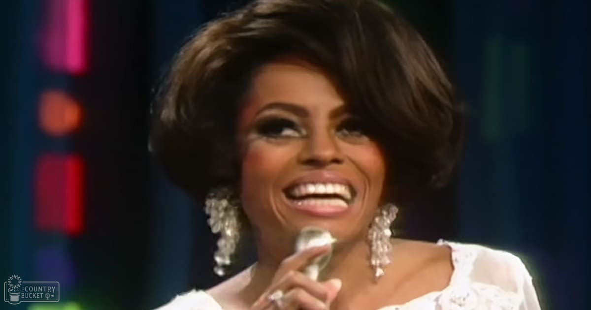 An adult female in a performance setting with large dangling earrings and rings, wearing white attire against red, pink, and blue stage lights.
