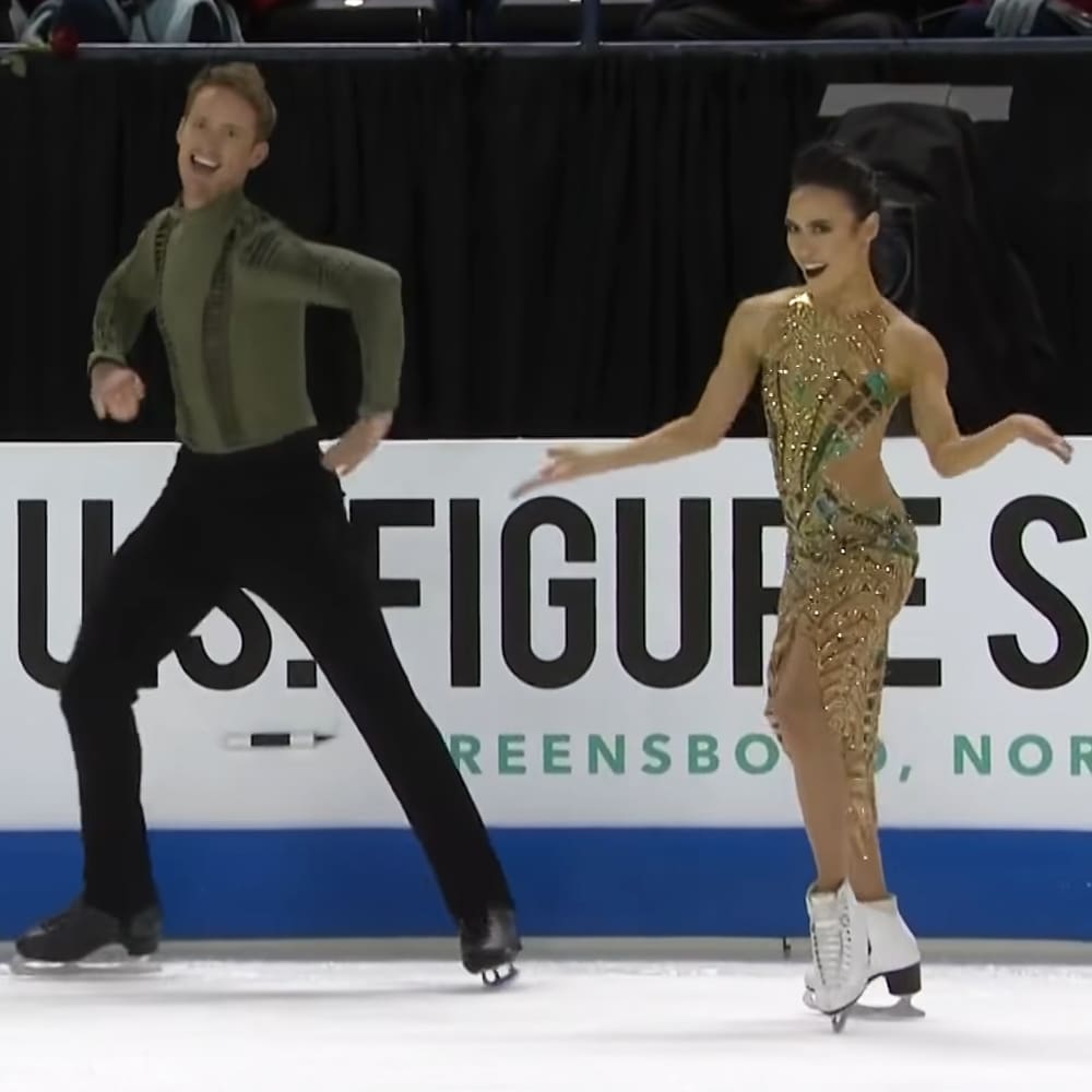 A young adult male figure skater in a green textured long-sleeved shirt and black pants stands with his female partner, wearing a gold sequined dress with an open back design at Greensboro's indoor ice skating rink. The setting is illuminated by natural daylight.