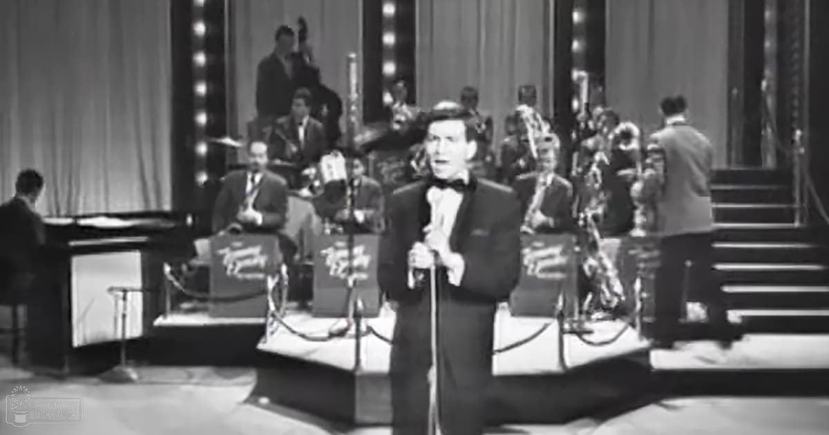 Orchestra members seated or standing on a stage, curtains visible, indoor setting with stage lighting at night.