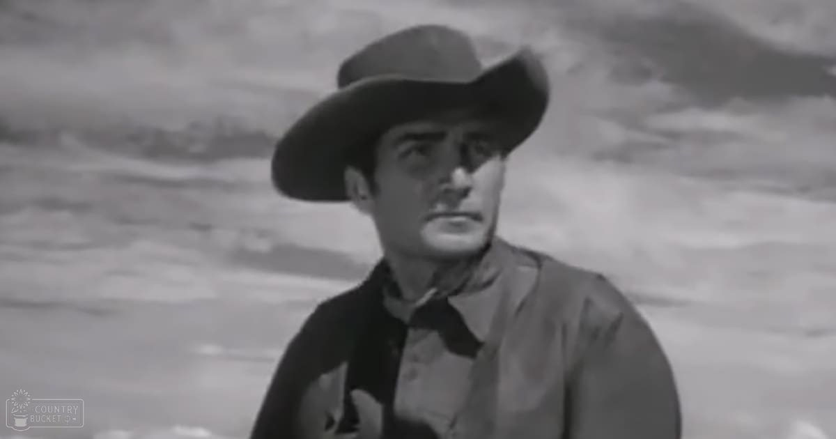 A man in a cowboy outfit stands against a desert landscape with mountains and sparse vegetation, wearing a felt hat, dark shirt, and long sleeves.