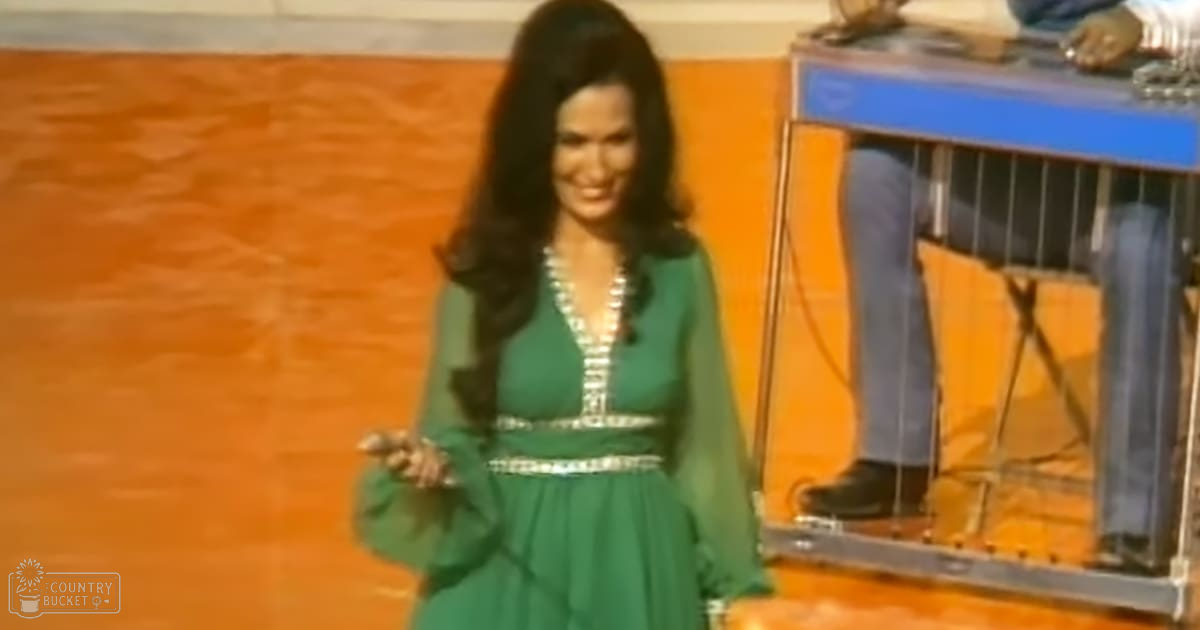 An adult female in a green dress with decorative elements, set against an orange wall and studio equipment, suggesting a television or radio broadcast environment.