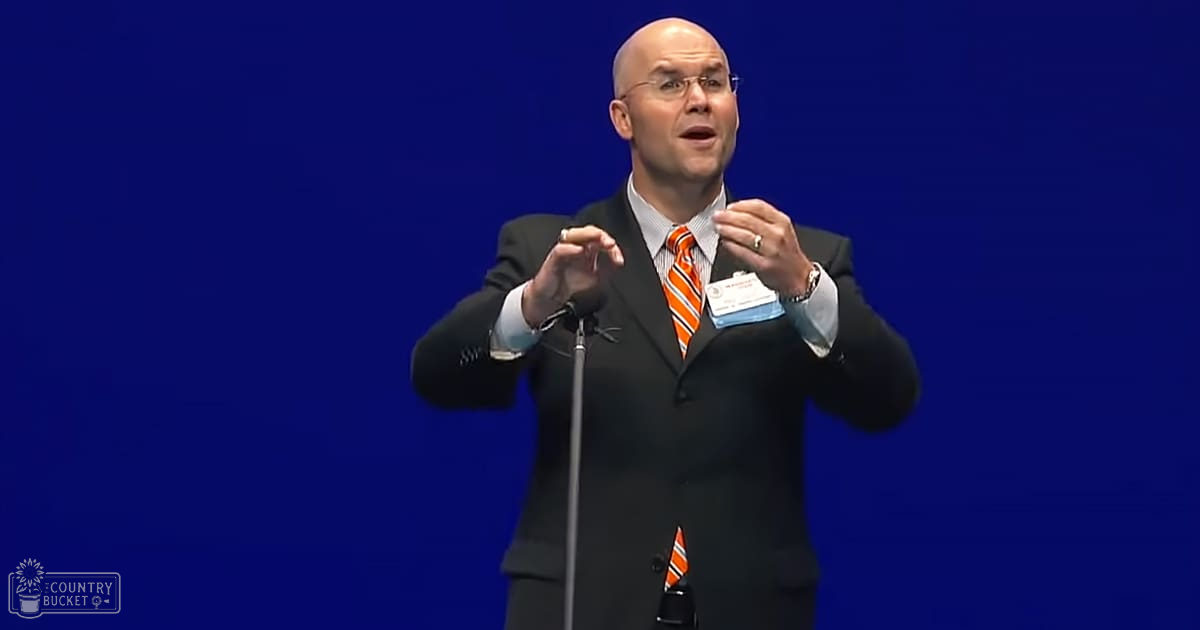 A middle-aged adult male wearing a dark suit, white shirt with an orange and white striped tie, giving a speech at an indoor event.