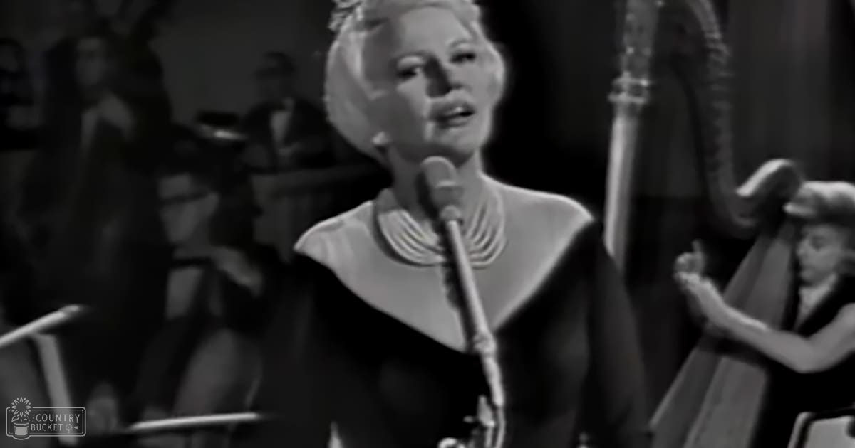 A woman singing into a microphone, accompanied by another individual who appears to be conducting or gesturing, both set against an indoor performance backdrop with stage lighting and musical instruments visible.