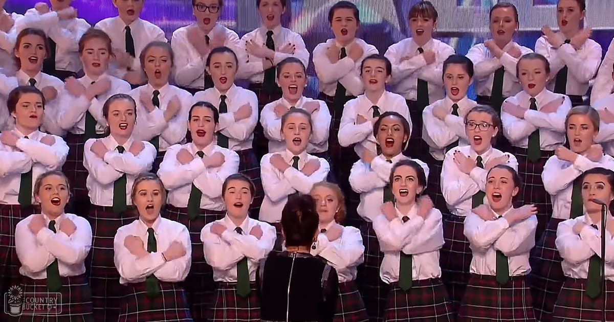 Teenage female choir performing at an indoor talent show or competition with white shirts, black ties, and red-and-green checkered knee-length skirts under blue stage lighting.