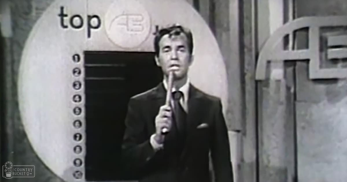 A young adult male in a dark suit with a pocket square stands in front of a "Top 40" sign, likely at a music event or radio station.