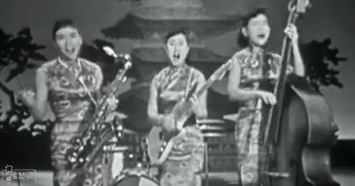 Musical performance scene with stylized Chinese architecture as backdrop; adult females wearing traditional patterned dresses perform on stage with a drum set and stringed instrument visible.