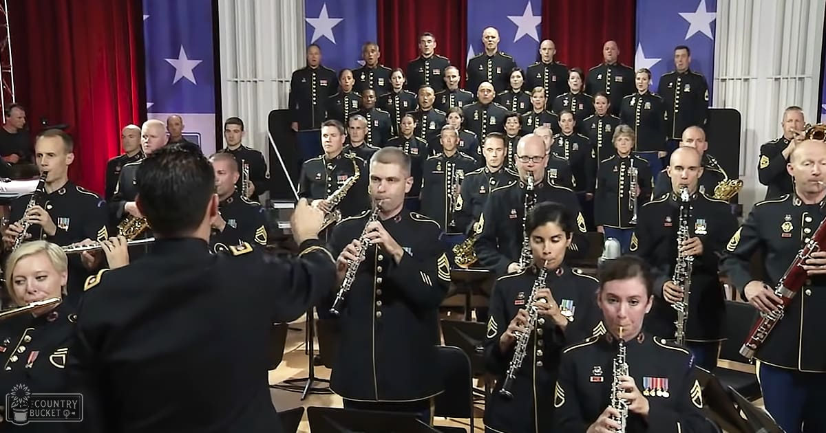Military orchestra performing formally indoors with red curtains and American flags behind them.