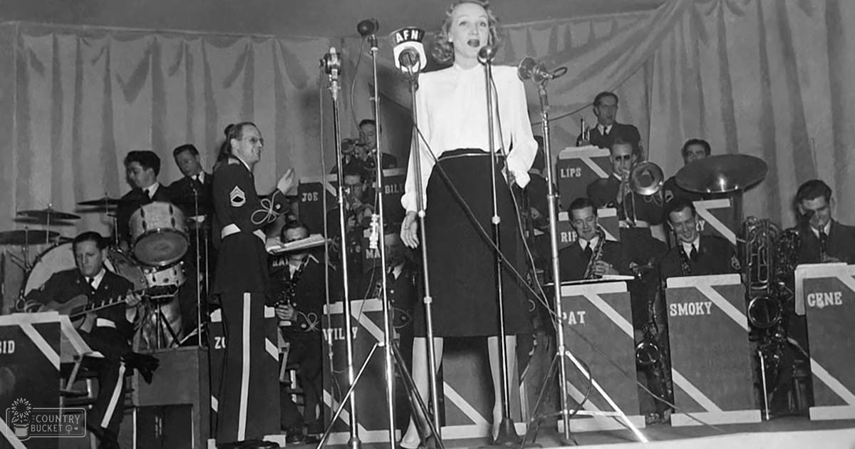 A woman singing in front of an orchestra at what seems to be a military base, with drum sets labeled 'JOE' and 'BIL'.