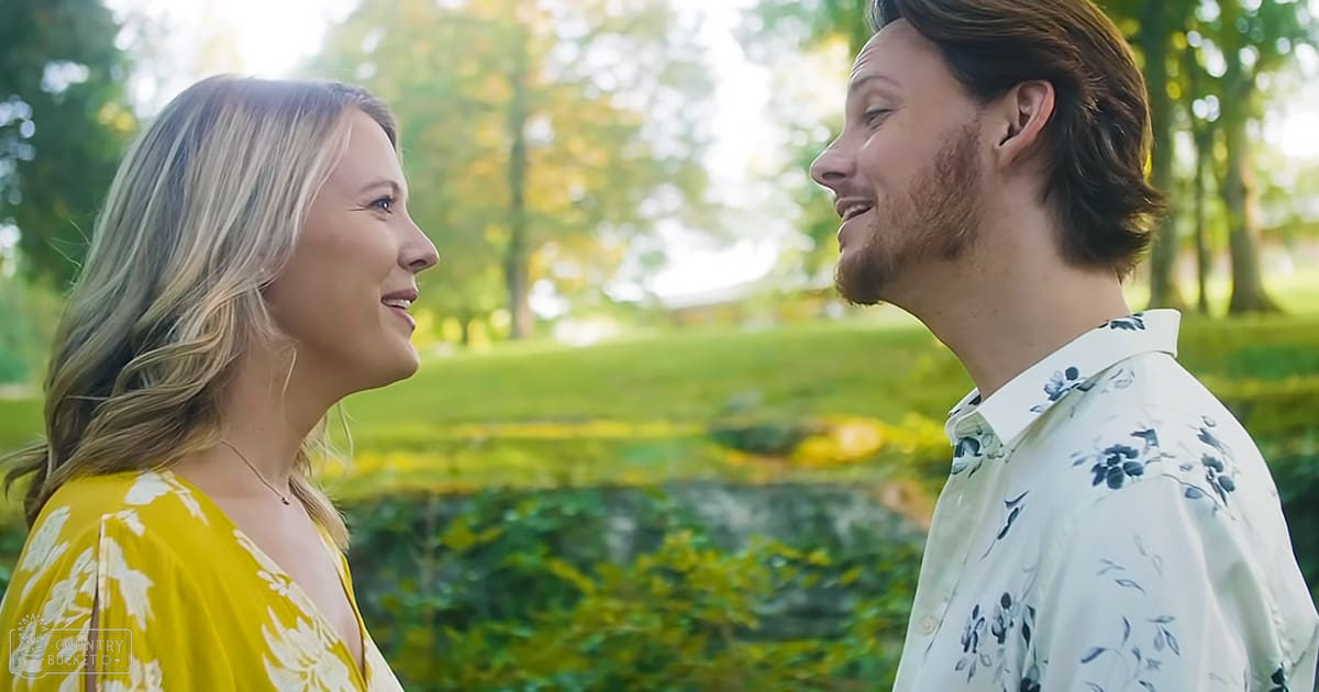 A young adult female wearing a yellow floral dress walks alongside a young adult male dressed in a white floral-patterned shirt in an outdoor park setting, with soft evening light and greenery.