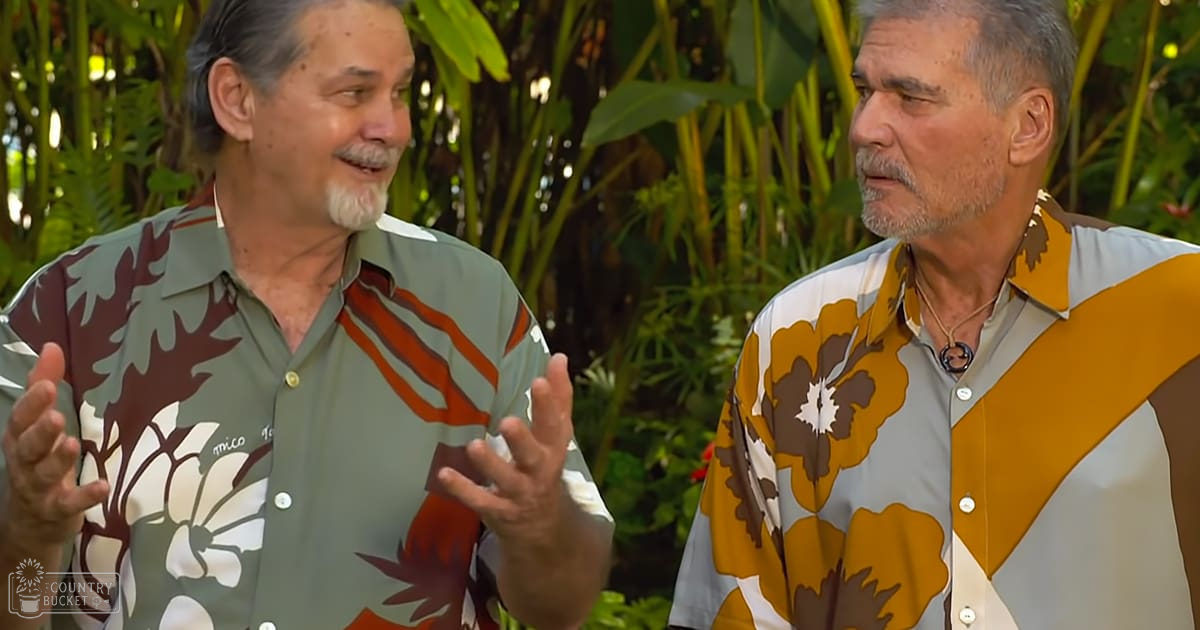 Two older adult males in Hawaiian shirts with floral patterns converse outdoors amidst greenery.