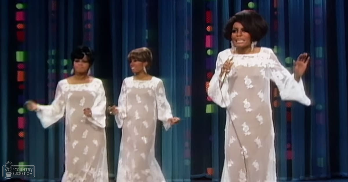 A performance scene with three adult females wearing bell-sleeved floral dresses, set against a blue curtain backdrop with colorful geometric designs. The lighting suggests it could be daytime or well-lit by artificial light typical of a performance venue.