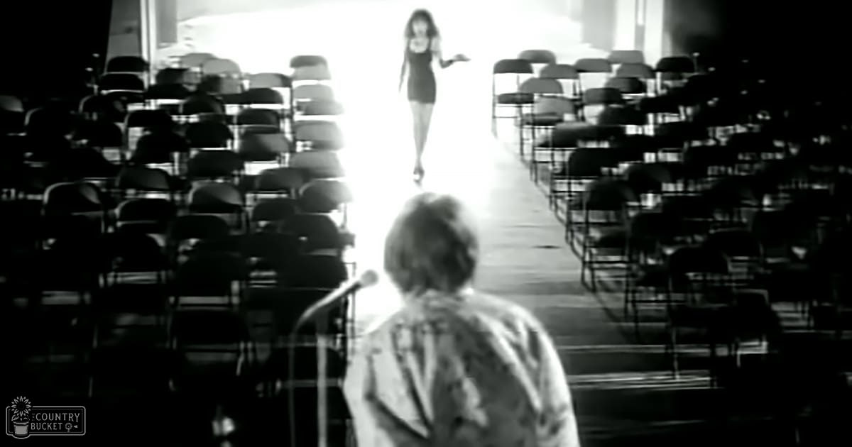 An adult female in a modern auditorium setting, wearing a black dress with a fitted cut from the mid-20th century; an adult male in the foreground wears a light-colored patterned jacket or shirt of similar era. The scene is indoors under artificial lighting and has rows of empty chairs.