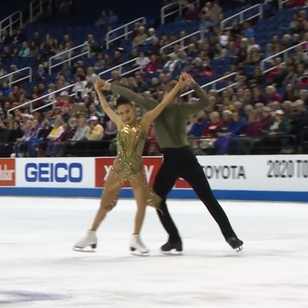 Two ice skaters performing at an indoor arena; female in a gold sequined dress, male in black pants and green shirt.