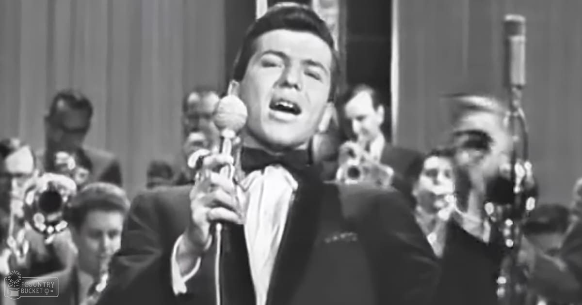 A male adult singer performing into a microphone, surrounded by band members in formal dark suits under bright indoor stage lighting.