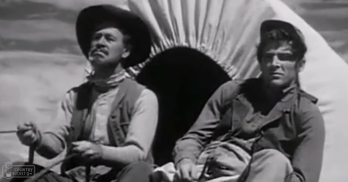 Two adult males in a Western setting wearing period-appropriate attire holding tools and standing near a canvas structure under natural daylight.