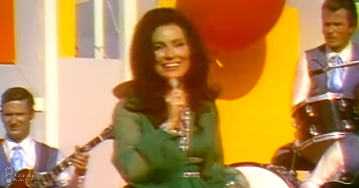A woman singing in a studio setting wearing a bright green dress with long sleeves, holding a microphone; surrounded by two men dressed formally, one in light blue and the other in white with a patterned tie.