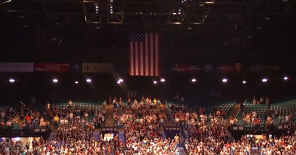 A crowded indoor arena during a sporting event with tiered seating, artificial lighting, and prominent American flag signage.