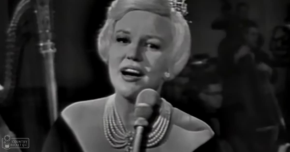 A middle-aged woman wearing a black high-collared garment with pearl jewelry sings into a microphone on stage, possibly from a film or performance.