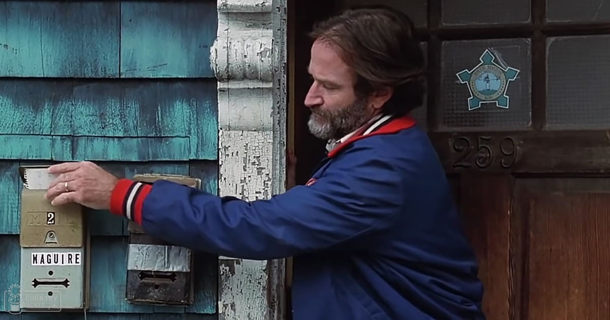 Middle-aged adult male in a blue jacket with red and white trim, standing outside a residential house labeled 'MAGUIRE' on the mailbox.