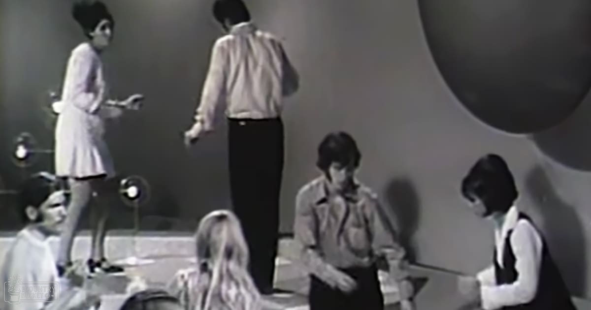 A performance scene with minimalistic indoor setting and controlled lighting; light-colored background, young adult female in white dress, adult male in cream shirt, and seated individuals in jackets and shirts.