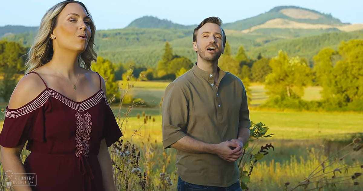 A young adult female in a maroon off-the-shoulder dress with intricate white embroidery stands beside an adult male wearing an olive-green button-up shirt, both in an open field with hills and greenery under warm late afternoon or early evening light.