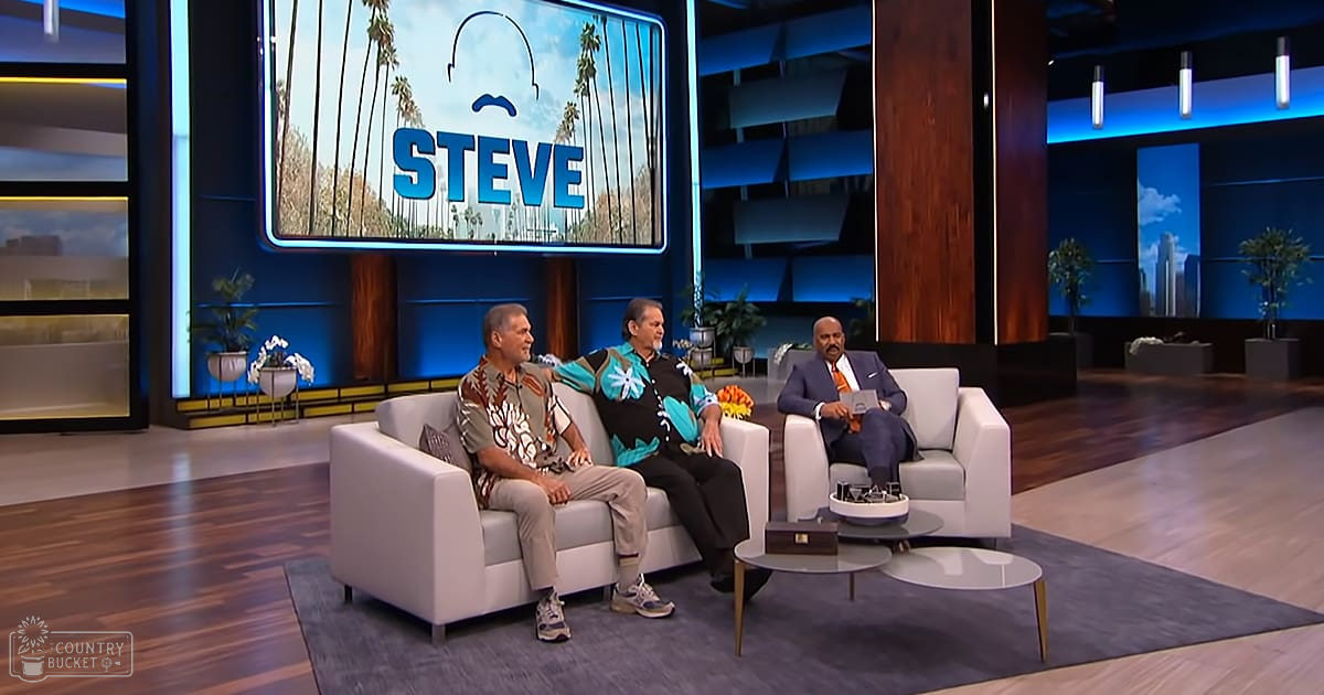 A talk show set in Los Angeles with two light-colored armchairs, a small round coffee table, decorative elements like potted plants, and a large screen displaying palm trees against blue skies.