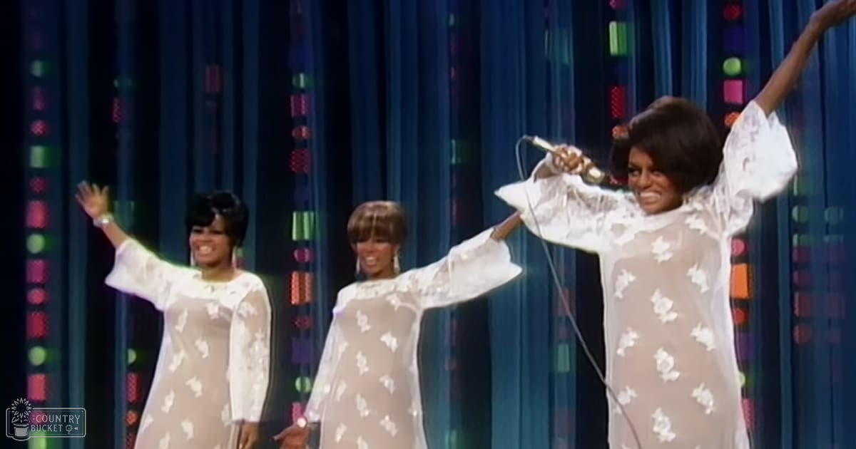 Young adult female performers wearing white floral dresses with bell sleeves, holding a microphone against a blue and colorful geometric-patterned curtain stage setting.