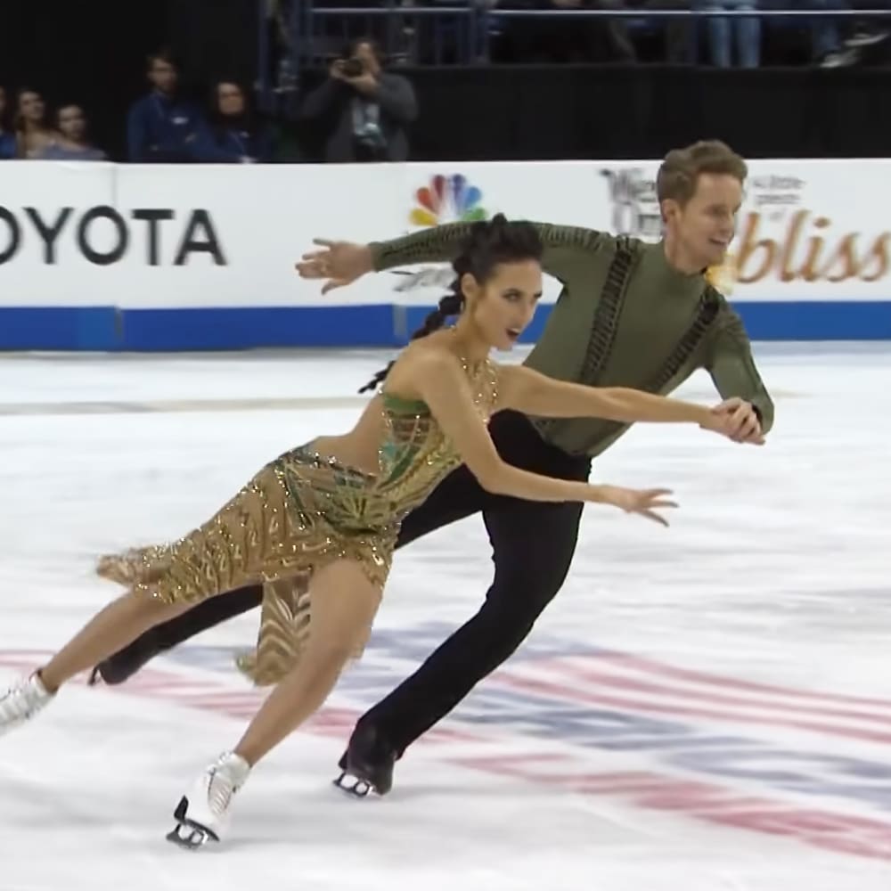 A figure skating pair performs at an indoor ice rink, surrounded by spectators on a sunny day.