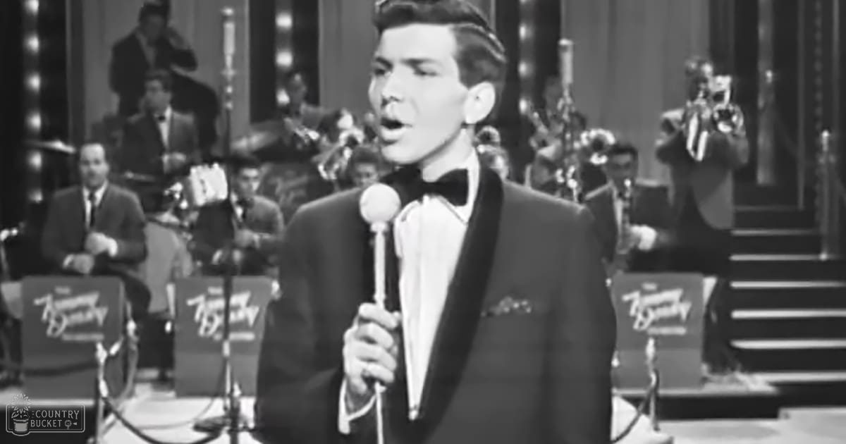 A young adult male singer performing on stage with an orchestra, dressed in a formal tuxedo, under indoor concert lighting.