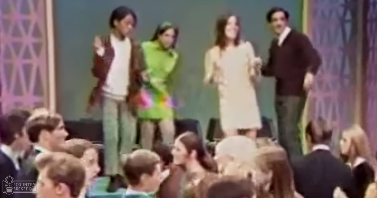 Group performance scene indoors with decorative panels and audience seating, possibly from a television show or stage production.